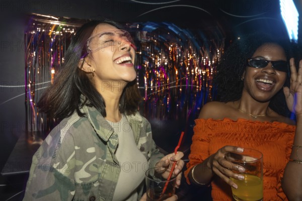 Two diverse young women enjoying a night out, laughing and dancing with drinks in hand amidst colorful club lights and festive decor, celebrating friendship and fun