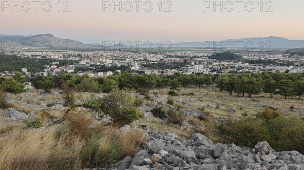 A city surrounded by nature with trees in the foreground and mountains in the background, illuminated by a warm sunset, looking out from a hill on the outskirts of Podgorica, Montenegro