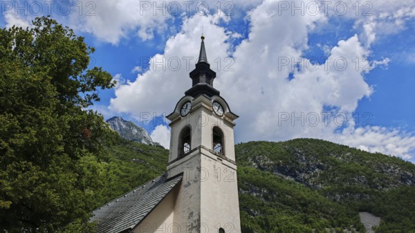 A church tower stands against a blue sky with clouds and a mountain backdrop, Slovenia