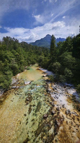 A clear river flows through a forest with Triglav mountains in the background under a blue sky, Soca Valley, Slovenia