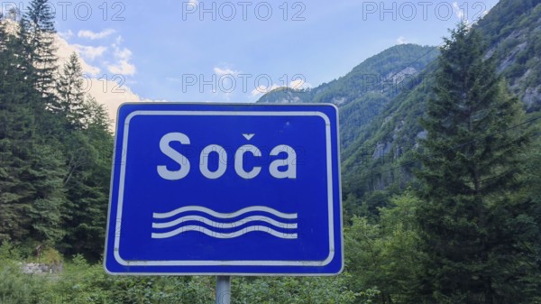 A blue road sign with the inscription 'Soca' in front of a mountainous forest landscape, Soca Valley, Slovenia