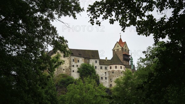 A historic castle rises behind thick greenery under a cloudy sky, Loket, Czech Republic