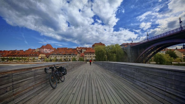 Wooden walkway with bicycle leading to a picturesque old town across the river under a blue sky with clouds, Maribor, Slovenia