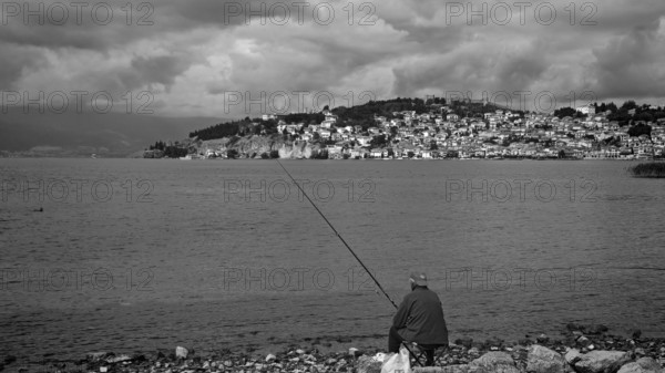 Black and white photo of a fisherman on a lake shore with a city in the background, Ohrid, North Macedonia
