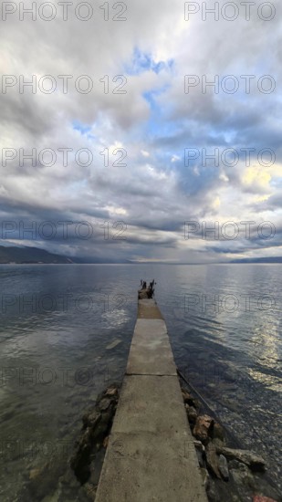 A long jetty leads into the calm sea under a cloudy sky that creates a peaceful atmosphere, Lake Ohrid, North Macedonia