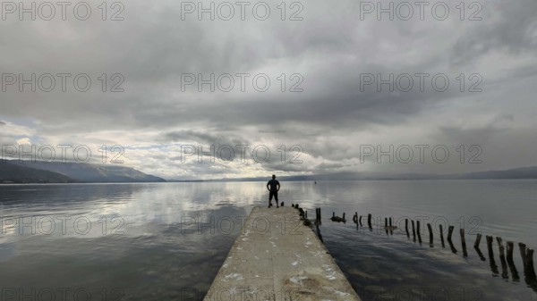 A man stands alone on a jetty that juts into a cloud-covered lake, Lake Ohrid, North Macedonia