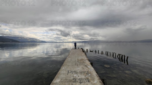 A man stands on a long jetty leading into a cloudy lake, Lake Ohrid, North Macedonia
