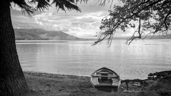 A boat on the shore of a calm lake surrounded by trees in black and white, Lake Ohrid, North Macedonia