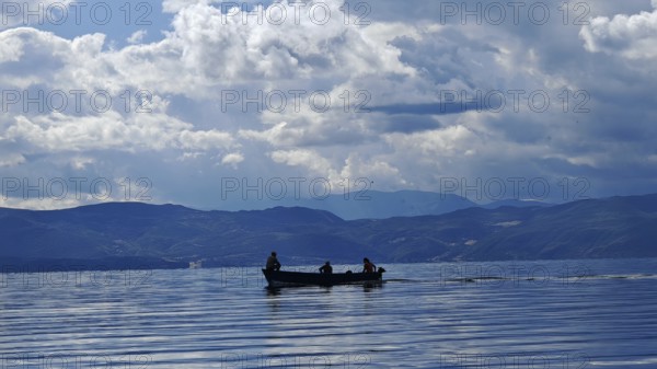 A small boat with silhouettes in a calm lake surrounded by mountains and dramatic cloud formations, Lake Ohrid, North Macedonia