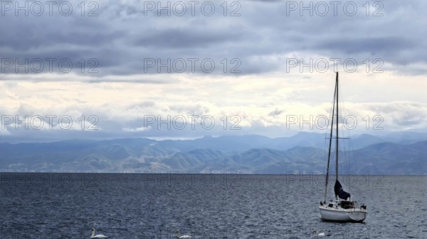 A sailboat on a calm sea with mountains in the background and cloudy sky, Lake Ohrid, North Macedonia