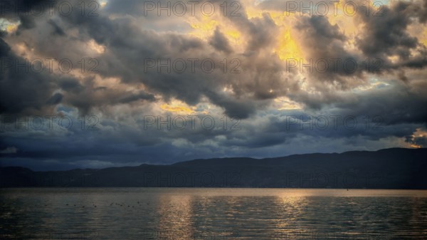 Dramatic clouds and golden light are reflected in the calm water of the lake during a sunset, Lake Ohrid, North Macedonia