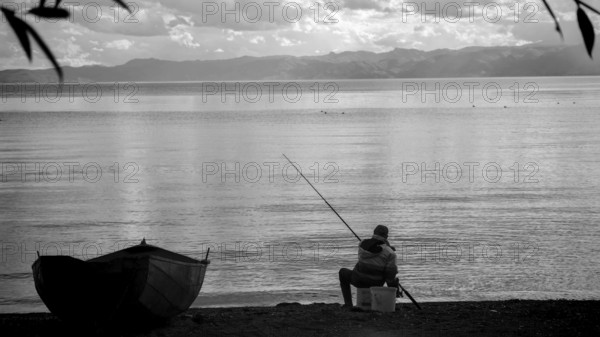 An fisherman sitting on the shore of a calm lake, with a small boat and a black and white aesthetic, Lake Ohrid, North Macedonia