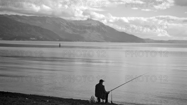 Angler sitting on quiet lakeside with views of mountains on horizon, Ohrid Lake, North Macedonia