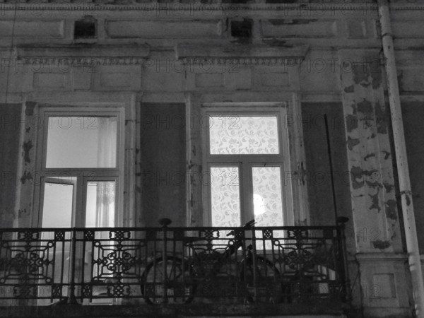 Worn façade at night with balcony on which a bicycle stands and illuminated windows in black and white, Poland
