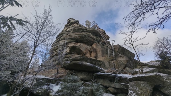 An impressive rock with a metal staircase, ladder, next to it, surrounded by snow-covered trees under a blue sky, Fichtelgebirge
