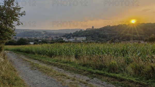 A trail leads along fields with a village in the background at sunset, Kronach with a view of Rosenberg Fortress behind a corn field, Upper Franconia