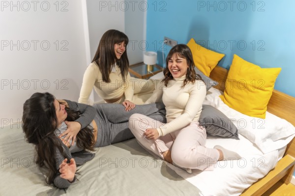 Three cheerful young women friends are enjoying a slumber party, laughing heartily while relaxing on a bed in a vibrant bedroom, sharing moments of friendship and happiness
