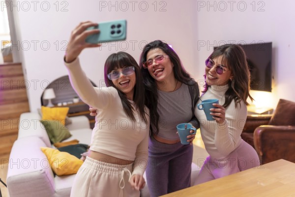 Group of happy young women friends wearing stylish sunglasses and holding drinks, capturing a fun selfie moment during an indoor party or casual gathering