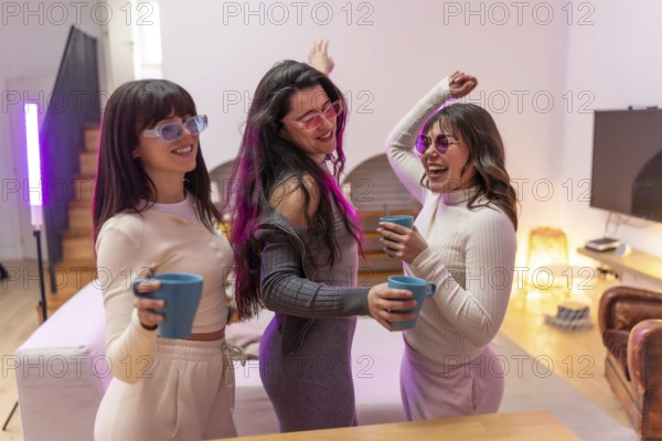 Three happy young women friends wearing sunglasses and casual clothes dancing, holding mugs, and laughing together while enjoying a home party with vibrant neon lights