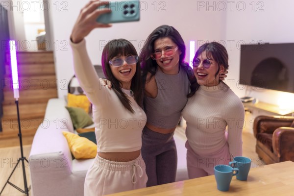 Three cheerful young women wearing colorful sunglasses gathering for a selfie in a modern living room with atmospheric lighting, enjoying a fun moment together