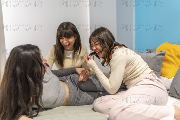 Three smiling young women friends relaxing on a bed, sharing a joyful moment and laughing together during a casual hangout at home, fostering friendship and connection
