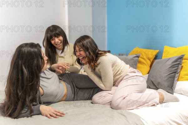 Three young women friends spending time together laughing and enjoying a slumber party on a bed during a leisure bedroom hangout, symbolizing friendship, togetherness, and happiness
