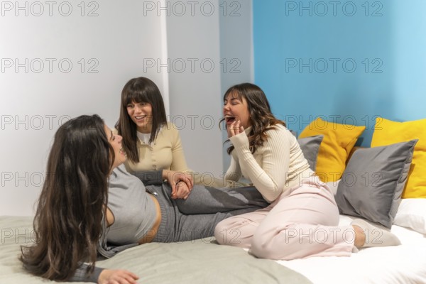 Three young women relaxing and enjoying a cheerful sleepover on a comfortable bed in a brightly decorated bedroom, sharing laughter and bonding during a casual gathering