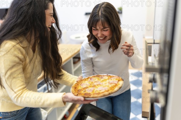 Two cheerful friends laugh in a cozy kitchen while presenting a freshly baked homemade pizza, enjoying cooking together and sharing a joyful, authentic moment of friendship and food