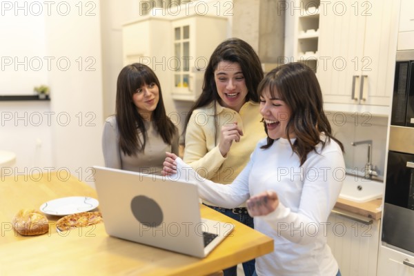 Group of female friends gathering in a modern kitchen, sharing an exciting moment while celebrating a victory, success, or positive news seen on a laptop screen