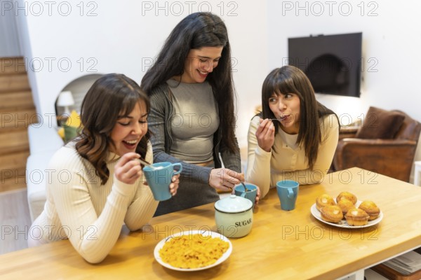 Three young adult women friends enjoying breakfast with muffins, coffee, and cereal, sharing smiles and laughter around a wooden table in a cozy home environment