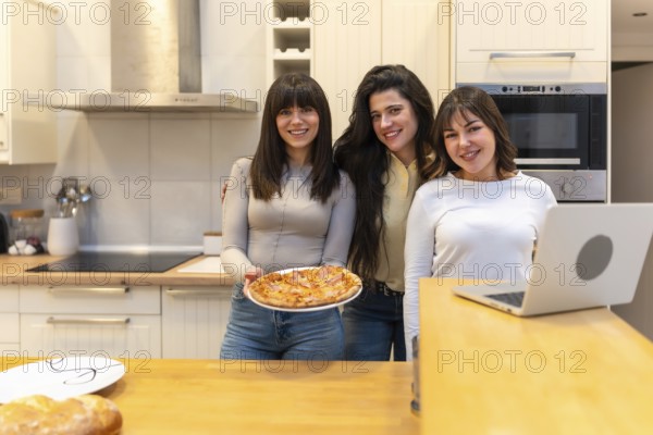 Three young women smiling and looking at camera while one is holding a freshly baked pizza in a modern kitchen, sharing a happy moment together at home