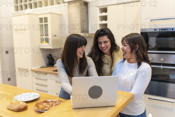 Three young women laughing together in a bright modern kitchen, watching a laptop while sharing pizza and bread, enjoying relaxed social time and friendly connection at home