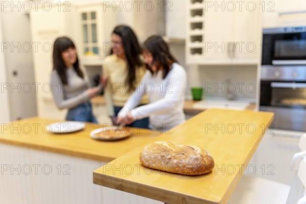 Loaf of artisan bread resting on a wooden kitchen counter while a group of three female friends enjoys preparing food in the blurred background, sharing a domestic moment