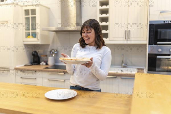 Young woman smiling in a modern kitchen while holding a freshly baked pizza on a plate, enjoying homemade cooking and domestic life, preparing a casual dinner with joy and pride