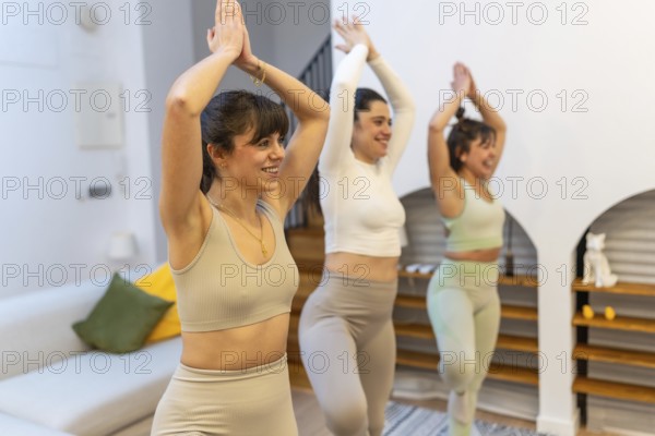 Three women smiling as they practice a balancing yoga pose together in a cozy living room, sharing mindfulness, fitness and friendship during a home workout session