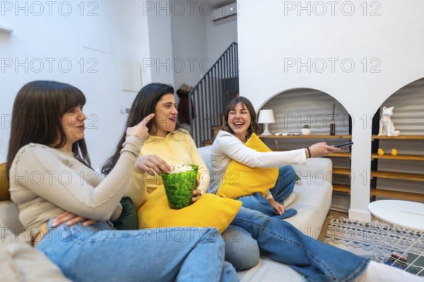 Three women on a sofa sharing popcorn, laughing and watching tv together in a cozy living room, enjoying a relaxed movie night and casual friendship at home