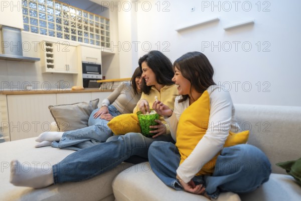 Group of diverse young women enjoying a relaxed evening on the couch at home, sharing laughter and popcorn while watching television together creating a bonding moment