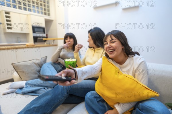 Three young women on a cozy sofa enjoying a movie night, laughing, sharing popcorn and relaxing together in a comfortable modern living room at home