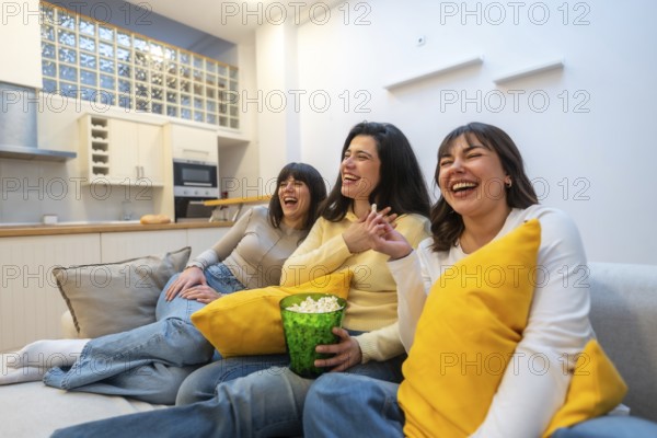 Three young adult female friends enjoying a relaxing movie night at home, laughing together, socializing, and eating popcorn while sitting comfortably on a sofa