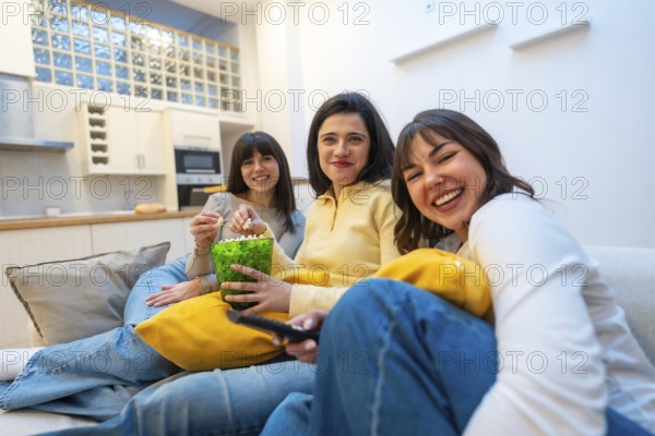 Three young women laughing and relaxed on a couch at home, enjoying a cozy movie night together with popcorn, streaming a film and bonding in casual clothing