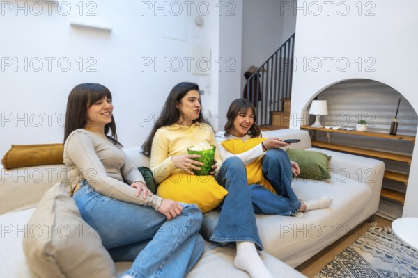 Group of happy young women friends relaxing on a comfortable couch at home, spending leisure time together, laughing and watching tv during a cozy movie night with popcorn