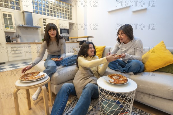 Three young women lounging on a sofa and floor, sharing pizza, laughing and chatting in a cozy apartment, enjoying relaxed weekend togetherness and friendship