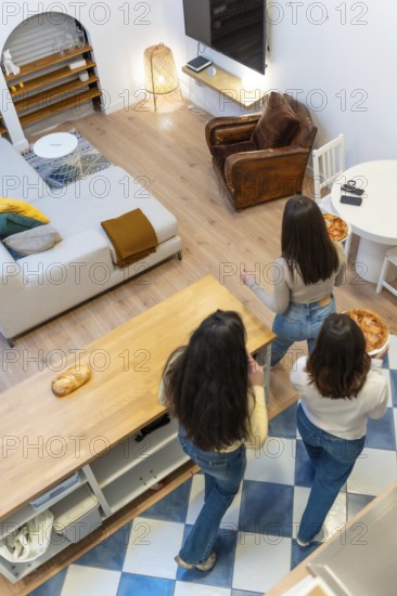Three young women gather in a stylish apartment living room, chatting and sharing pizza in a comfortable and friendly atmosphere that highlights togetherness and warmth