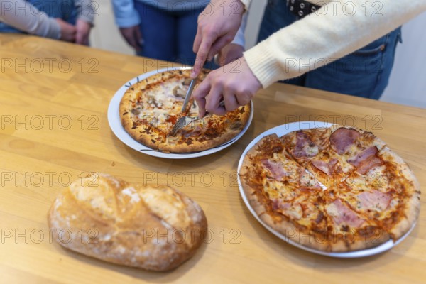 Person's hands using a knife and fork to cut a hot pizza into slices, ready for sharing a casual meal with friends or family gathered around a wooden table with another whole pizza and fresh bread