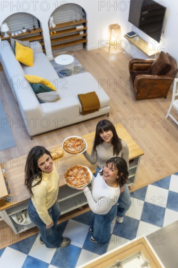 Three cheerful young women standing in an open plan apartment, holding pizzas and looking up while smiling, showing friendship and enjoying domestic leisure