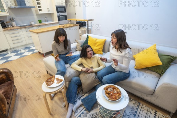 Three young women gather on a comfortable sofa in a modern living room, sharing pizza slices and engaging in relaxed conversation during a casual dinner at home