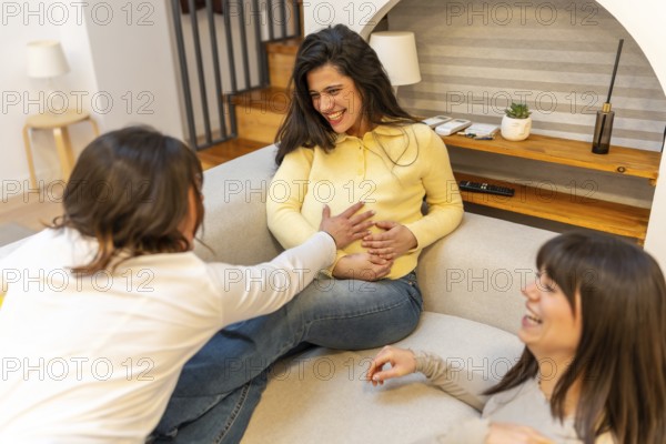 Three cheerful women sharing a moment of joy and friendship at home, one friend touching the expectant mother's baby bump as they share laughter and anticipation for new motherhood