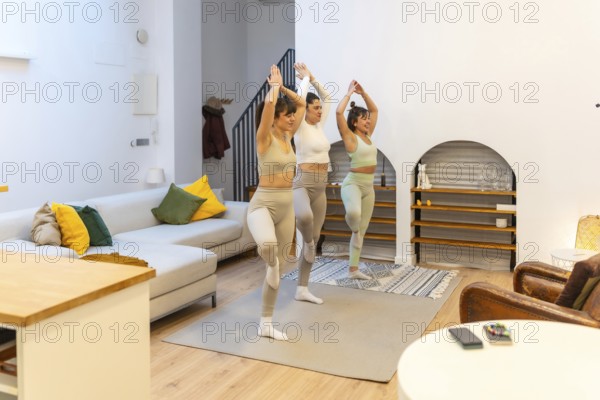 Three young women practice tree pose together on yoga mats in a modern apartment living room, balancing on one leg for fitness, mindfulness, and friendly home workout harmony