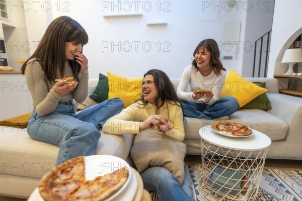 Three young women relaxing on a couch and floor, sharing pizza slices during a casual gathering, enjoying friendship and candid moments of laughter indoors