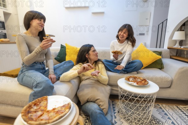 Three young women laughing and talking while eating pizza slices on a sofa in a modern living room, sharing a leisurely meal and friendship during a relaxed gathering
